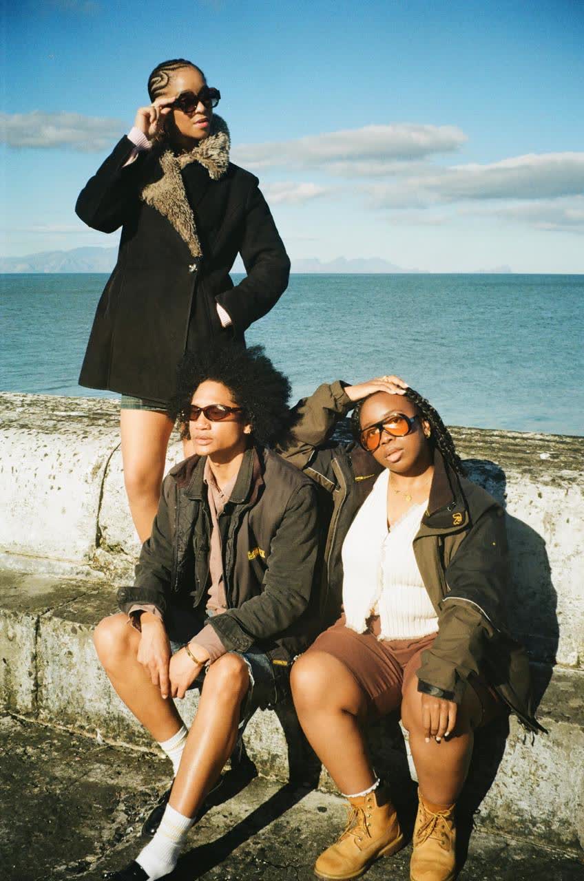 Three friends in thrifted jackets and shorts posed on a sea wall